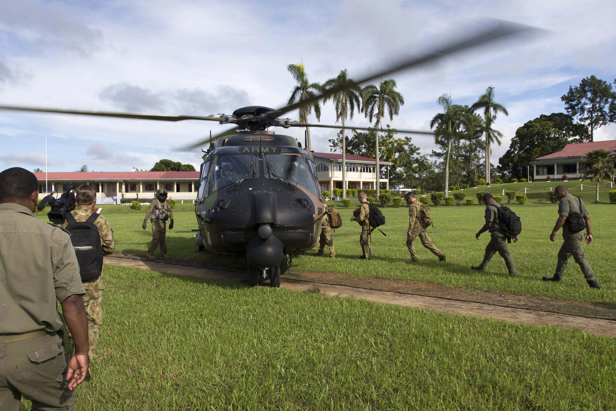 Soliders walking across green grass approaching a black helicopter marked army; colonial-style buildings and palm trees in the background