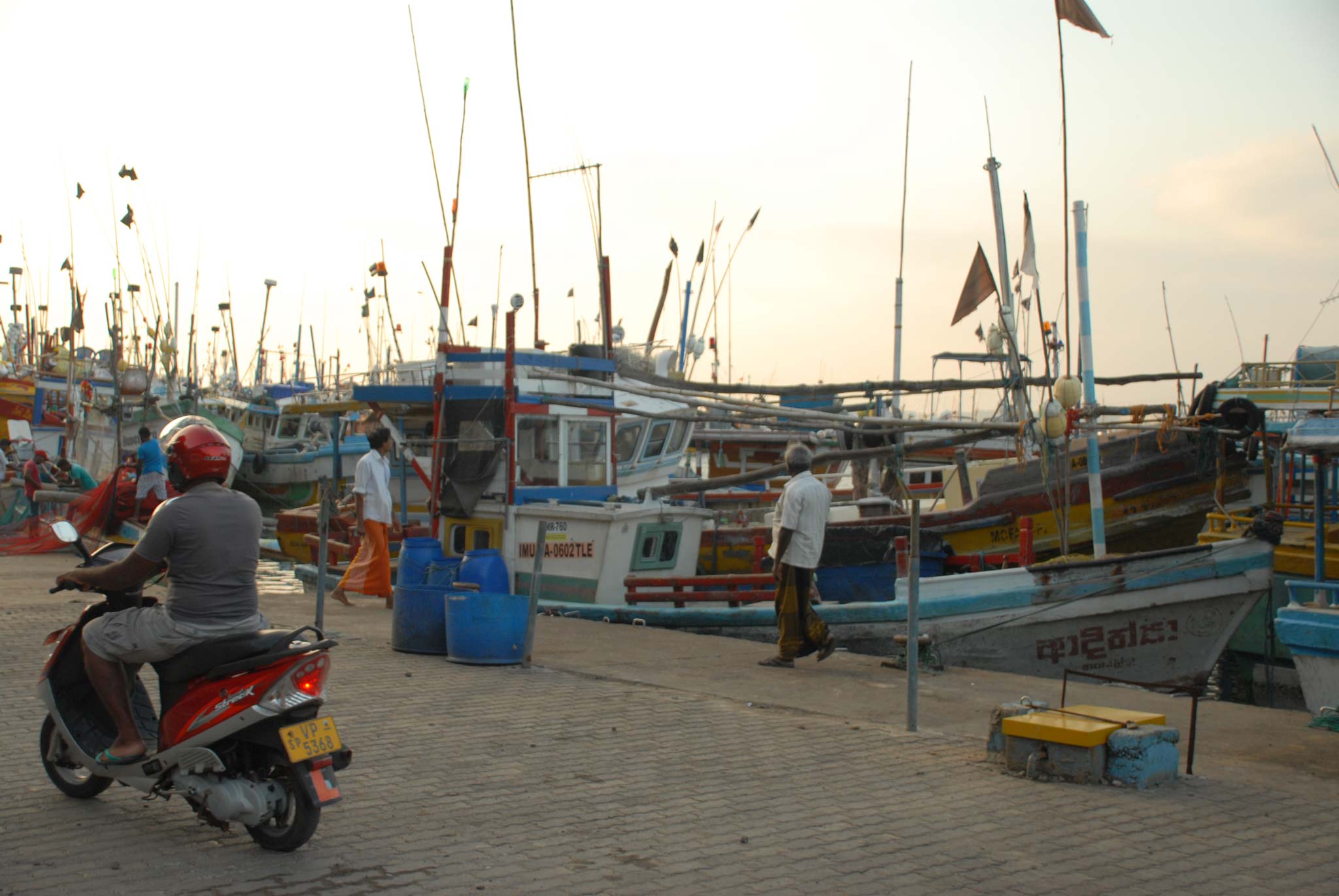 Port scene, dusk, with many wooden fishing boats, two men and a motorbike in foreground 