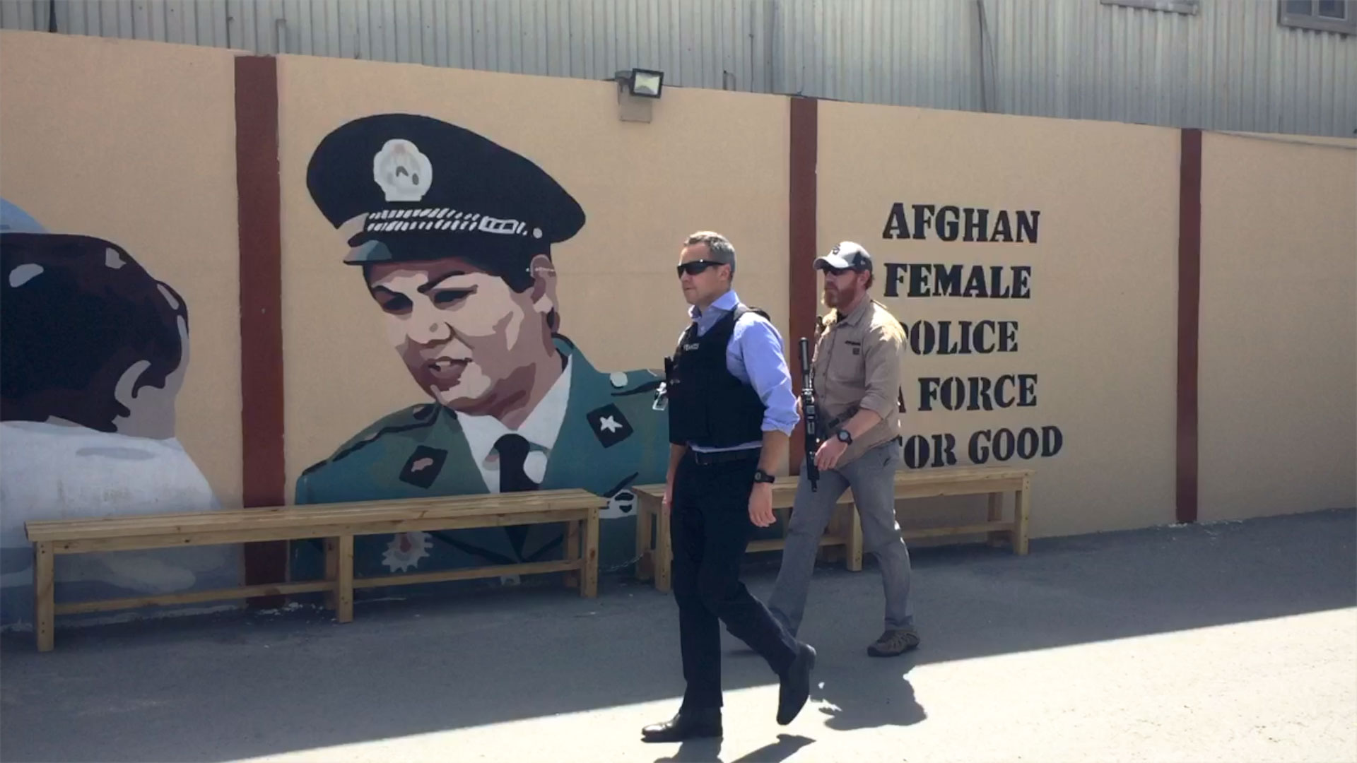 Richard Feakes, wearing flak jacket walking with a solider, in front of a mural of a female police officer and (partially obscured) wording 'Afghan female police is a force for good'