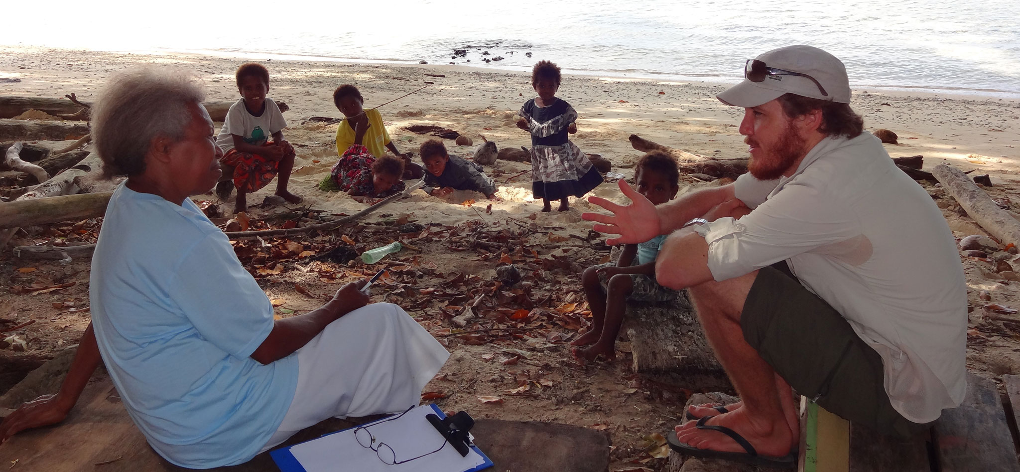 Tim Baird seated on a log talking with an older woman with children and the sea in the background, Papua New Guinea