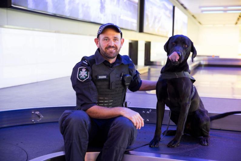 Mathew Blow sitting on airport conveyor belt with black labrador sniffer dog Leo