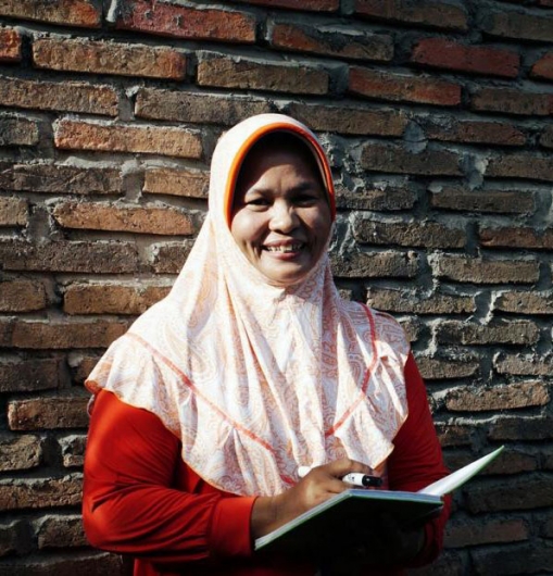 Syamsiah, pictured form the waist up, holding a notebook and pen, near a poster with writing in Bahasa Indonesia