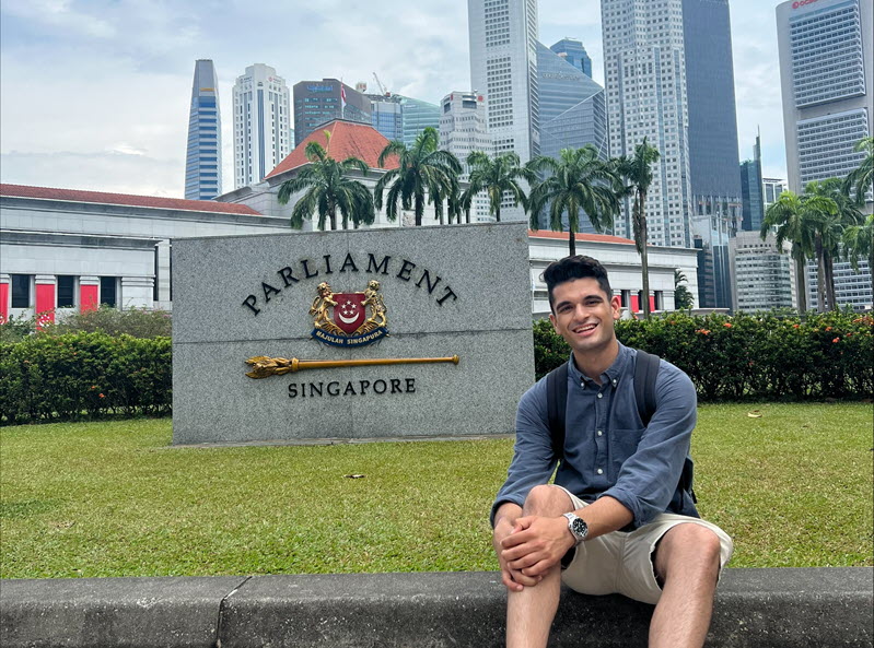 Ricky sitting on the grass in front a Singapore Parliament sign with the city skyline in the background.