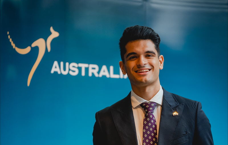 Ricky standing in front of an 'Australia' nation brand banner, dressed in a dark blue suit, white business shirt, and a maroon tie with white chevron pattern. 