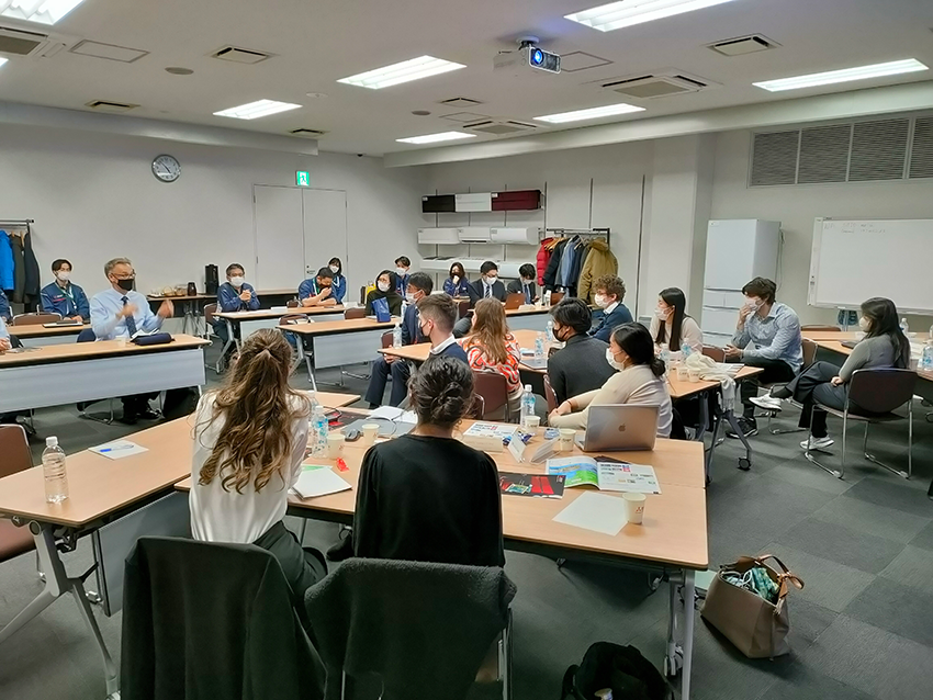 A room full of participants sitting at tables with notes and pens, listening to the lecturer