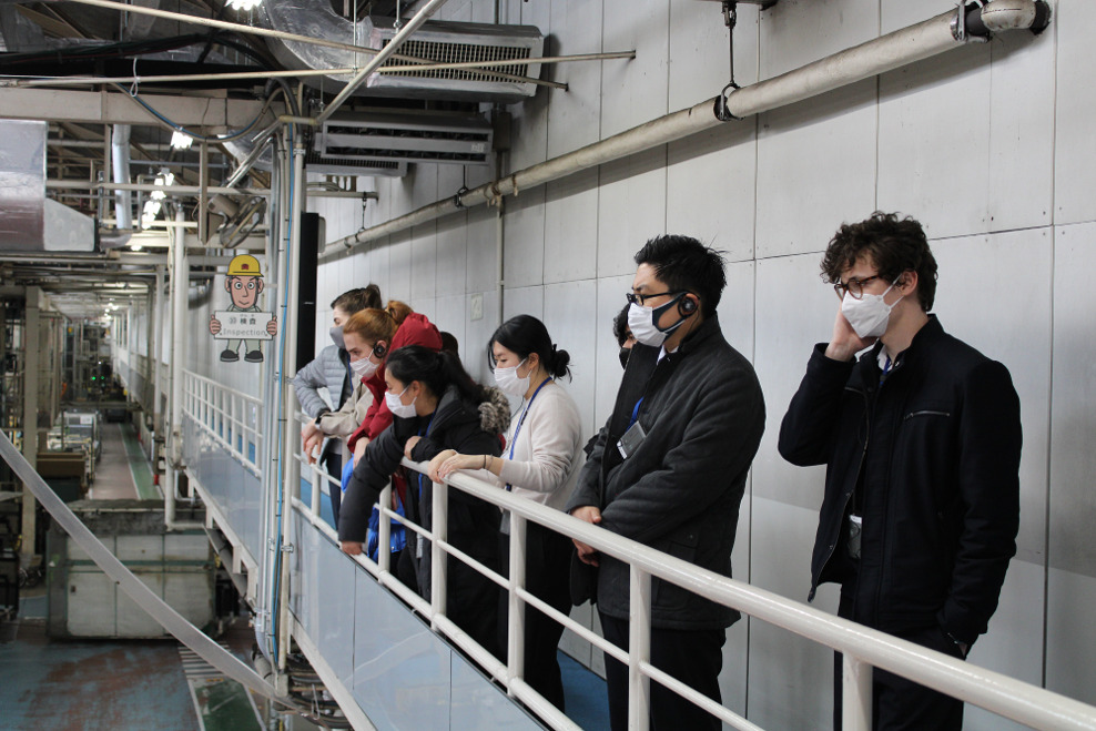 Several people wearing masks, leaning over a rail inspecting a workshop on the platform below