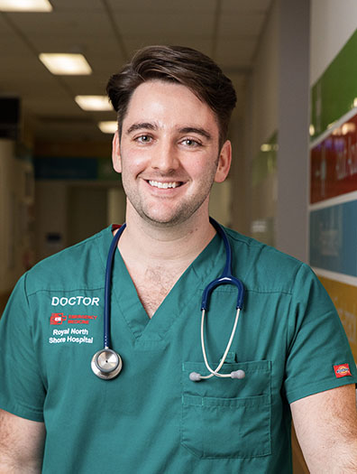 Man standing in a hallway in hospital scrubs with a stethoscope around his neck.