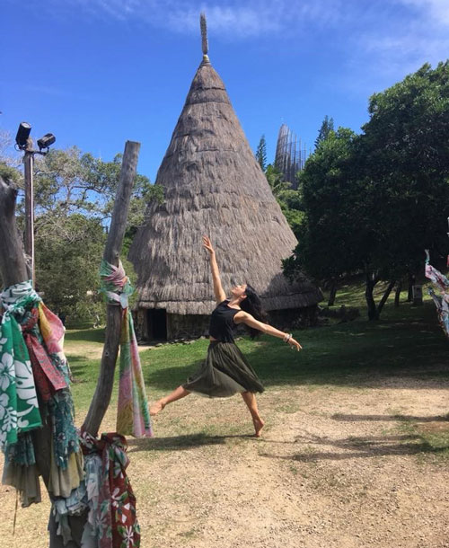 Ella performing a dance move in the local community with a tall hut in the background.