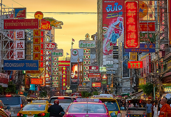 A busy Bangkok street with tuk-tuks, cars and pedestrians and Thai billboards.