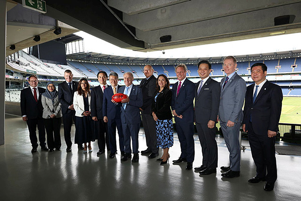 A group of people dressed in formal business attire standing in a row inside a large stadium concourse. The stadium seating and field are visible in the background through an open area. One person in the center is holding a bright red Australian rules football. The setting appears to be a professional or corporate event at a sports venue.
