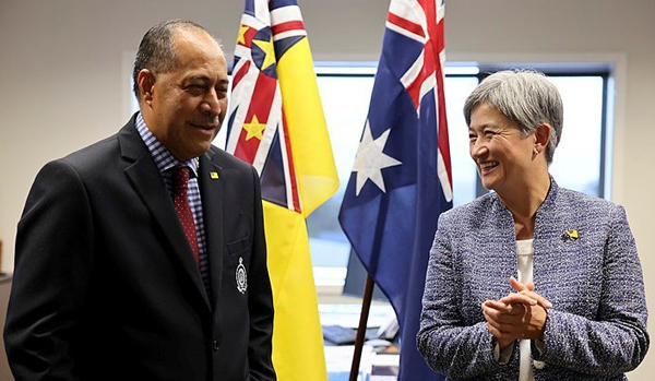 Two people standing and talking indoors, with the flags of Niue and Australia displayed behind them.