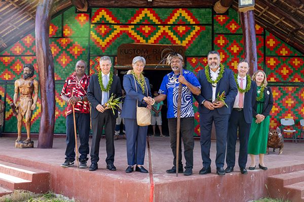 Eight people in formal attire wearing green garlands stand in front of a building labeled 'KUSTOM BUNGALOW.' One person shakes hands with another holding a staff, while a wooden statue is visible on the left. The building features colorful, patterned walls, suggesting a ceremonial or cultural event.