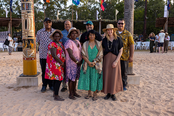 ight people in colorful attire stand on a sandy area with trees, flags, and decorated poles in the background. The scene appears to be part of a cultural or community event, with chairs and other attendees visible behind them