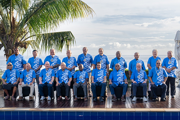 Eighteen people in matching blue patterned shirts pose on a wooden deck with a tropical ocean view and palm tree in the background. They are arranged in two rows, suggesting a formal group photo at an outdoor event.