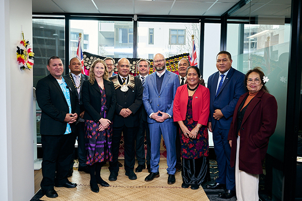 Eleven people in business and traditional attire pose for a group photo in a formal indoor setting with large windows, flags, and decorative elements in the background, suggesting an official or ceremonial event.