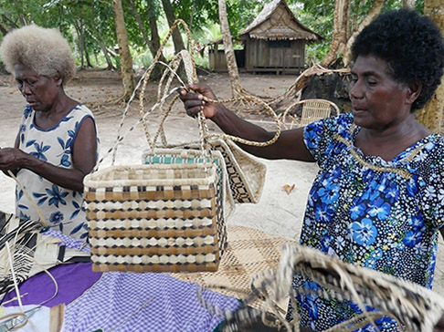 2 women at table outside, 1 holding up woven baskets