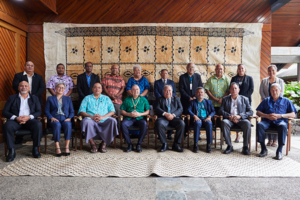 Group photo of delegates seated and standing in two rows in front of a large patterned tapestry, inside a timber-roofed venue. Participants wear formal and patterned attire, arranged on chairs and standing behind.