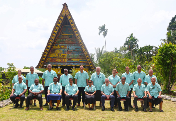Group of 20 people in matching light aqua shirts, seated and standing in front of a colourful traditional building surrounded by tropical gardens.