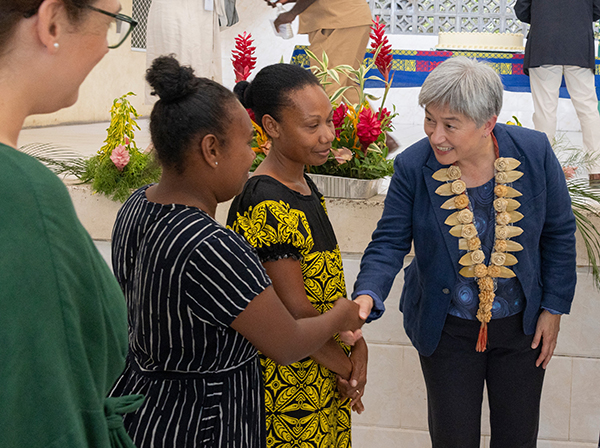 Person in a blue jacket and large necklace shakes hands with two people wearing patterned clothing in a courtyard with plants and flowers, while another person observes nearby.
