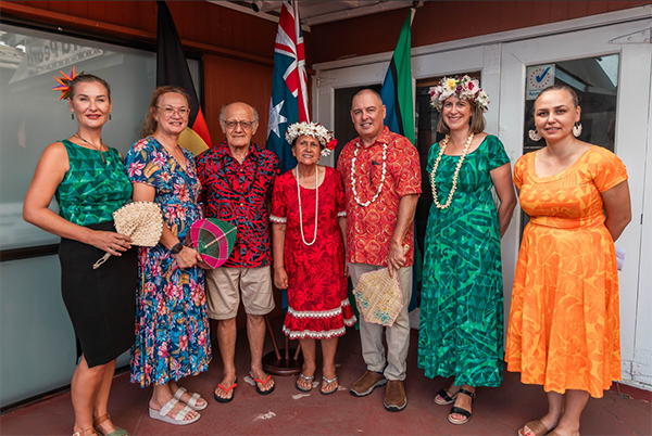 Six people standing in front of a building, wearing colorful traditional clothing, floral headpieces, and leis, with flags and a sign in the background, suggesting a cultural or festive event.