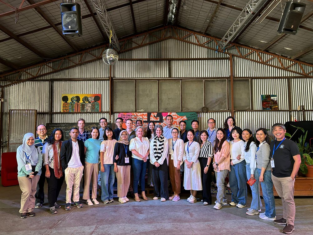 Photograph of a large, diverse group posing together inside an industrial event hall, likely documenting a team event. Corrugated-metal walls, hanging speakers, disco ball, colorful banners, plants, and casual-to-business attire surround participants arranged in a single row.