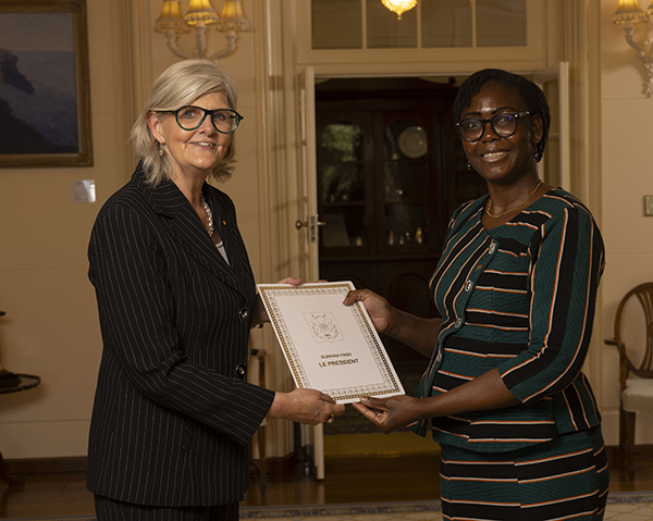 Two people stand indoors exchanging a formal certificate. One person wears a dark pinstripe suit, and the other wears a striped dress. A cabinet and decorative lighting are visible in the background.