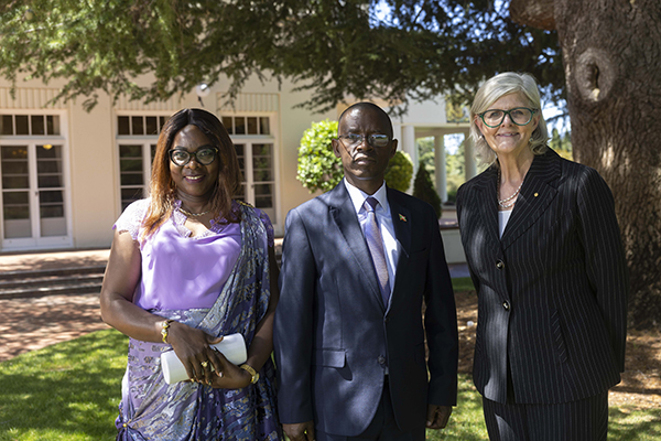 Three people stand outside near a large tree and a light-colored building. One person wears a purple and silver sari-style outfit and holds a clutch. Another person wears a dark suit with a tie, and the third person wears a dark pinstripe suit.
