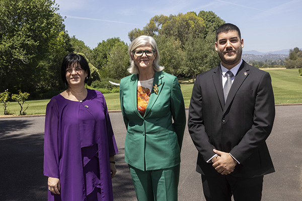 Three individuals are standing outdoors on a paved area with lush green grass and trees in the background. One person is wearing a green suit with an orange patterned top, another is in a purple outfit, and the third is in a dark suit with a tie.