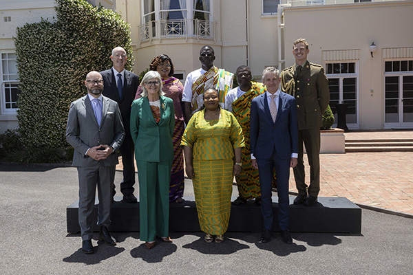 A group of nine individuals is posed for a formal photo outdoors in front of a cream-colored building. They are standing on a black platform. Attire includes suits and traditional patterned garments in vibrant colors such as green and yellow.