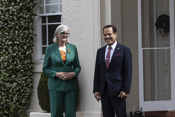 wo individuals are standing outside near a cream-colored building with a white-framed door and window. One person is wearing a green suit with an orange patterned top, and the other is in a dark suit with a tie. A leafy plant is visible on the left side.