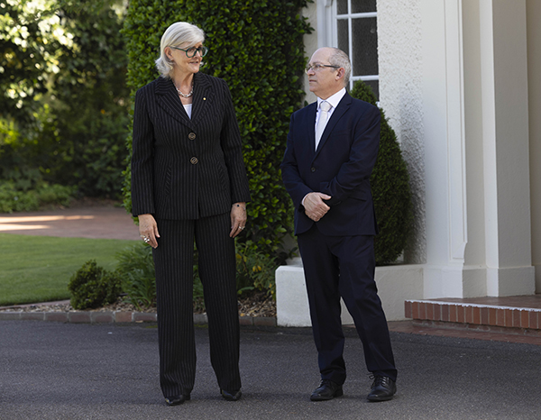 Two people stand outside a light-colored building with manicured greenery behind them. One person wears a dark pinstripe suit, and the other wears a dark suit with a white shirt. They are positioned on a paved area near the entrance steps.