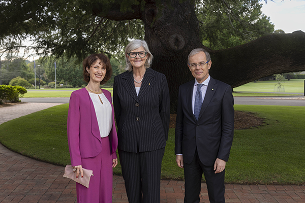 Three people stand outdoors on a brick path in front of a large tree. One person wears a bright pink suit and holds a light-colored clutch. Another person wears a dark pinstripe suit, and the third person wears a dark suit with a tie. A lawn and scattered trees are visible in the background.