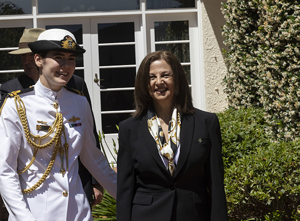 Two individuals are standing outdoors near a cream-colored building with white-framed windows and greenery. One person is wearing a white military-style uniform with gold cords and medals, and the other is in a black suit with a patterned scarf.