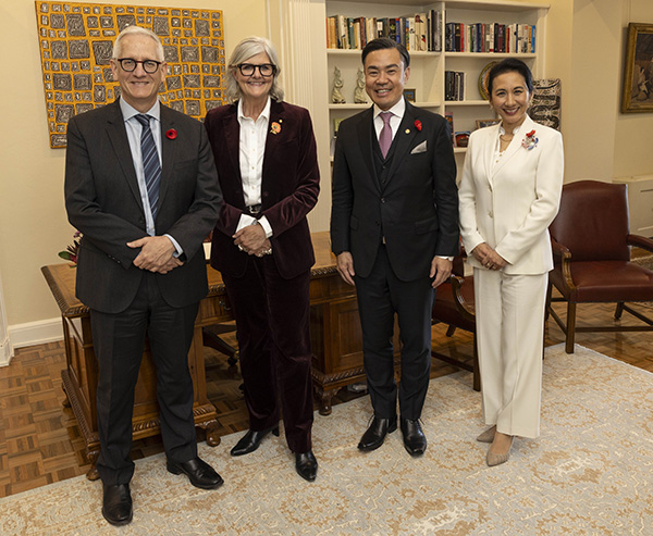 Four individuals are standing indoors in a room with wooden flooring, bookshelves, and framed artwork. They are dressed in formal attire, including dark suits and a white suit. Red poppy pins are visible on their clothing.