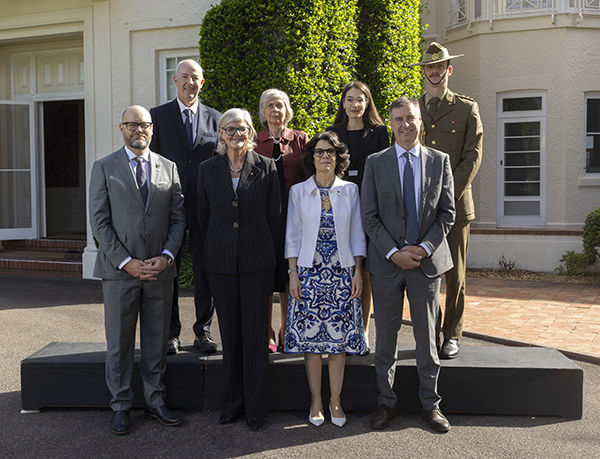 A group of eight people pose for a formal photograph outside a light-colored building. They stand on a small platform. The individuals wear a mix of suits, a patterned dress with a white jacket, and one person in a military uniform. Shrubs and the building’s facade are visible behind them.