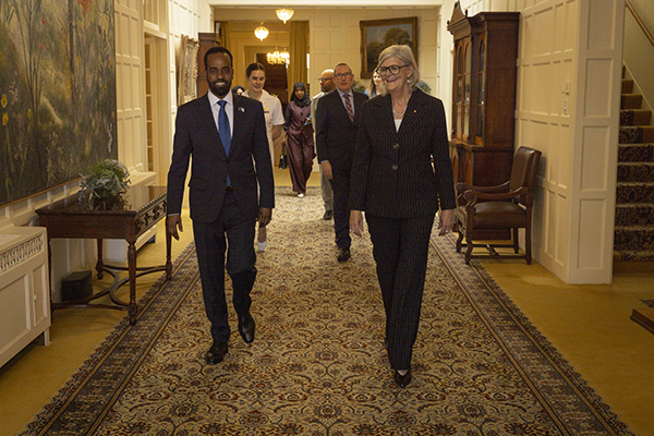 A group walks down a carpeted hallway inside an elegant building. The two people at the front wear formal suits and walk side by side. Several others follow behind them. The hallway features framed artwork, wooden furniture, and warm lighting.