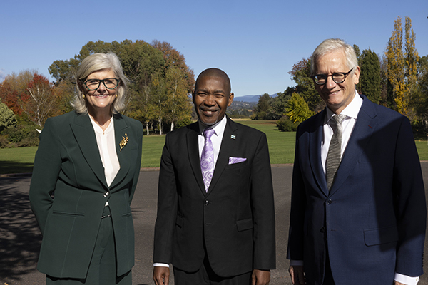 Three people in formal suits stand outdoors on a paved path with a large lawn, trees, and clear blue sky in the background.