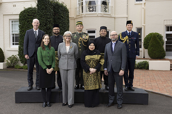 Nine people pose outdoors in front of a building, standing on a small platform, wearing a mix of formal suits, uniforms and traditional clothing.