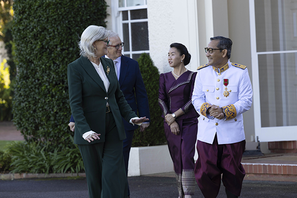 Four people in formal and ceremonial attire walk and talk outside a white building with tall windows and greenery.