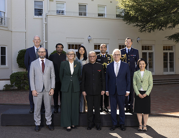 A group of formally dressed people pose in two rows in front of a light‑colored building in a courtyard setting.