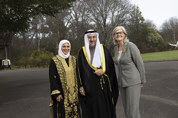 Three people stand together outdoors on a paved area with trees in the background, wearing formal suits and traditional attire.