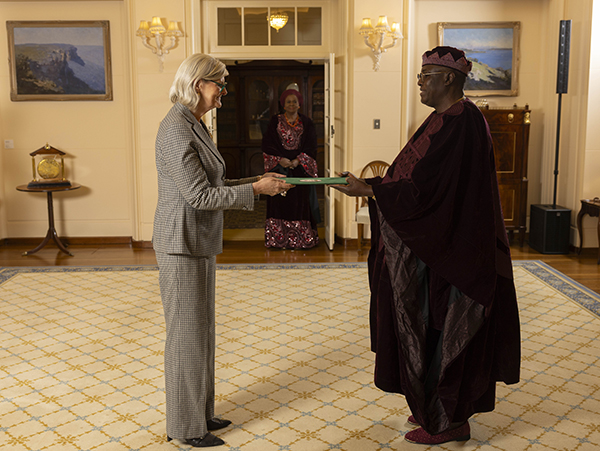Two people stand facing each other in a formal room, holding a document folder between them, with a third person seated in the background near a cabinet and framed artwork on the walls.