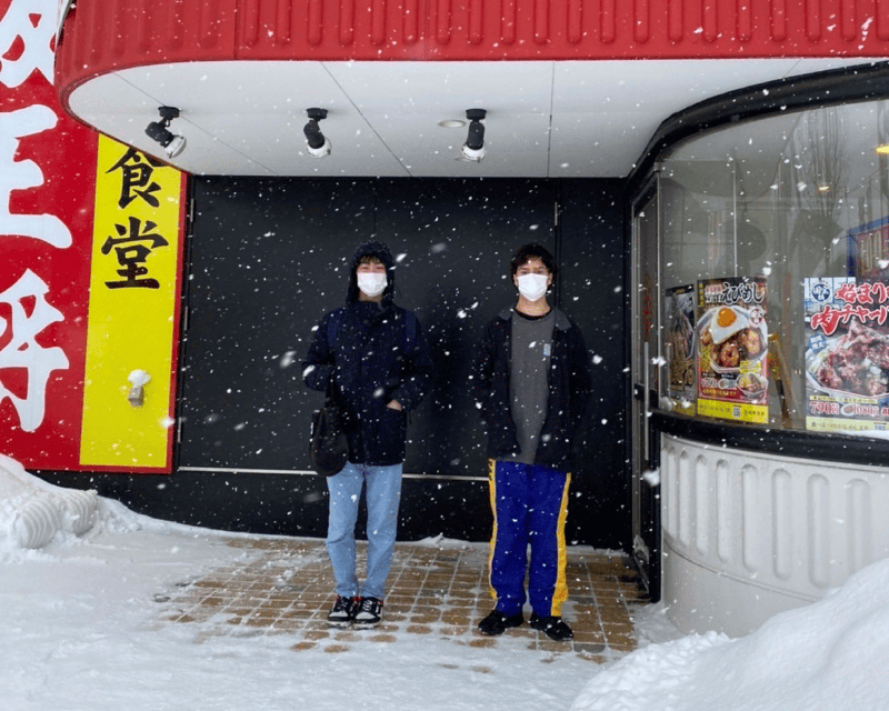 Two people standing in a doorway, sheltering from snow. Japanese signage on the wall in the background