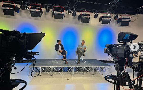 Television studio set with two speakers in conversation on a raised stage, filmed by broadcast cameras and illuminated by blue and yellow backdrop lighting.