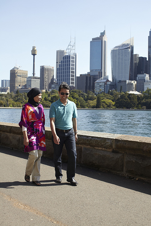 A man and a woman walking next to the water in front of the Sydney cityscape.