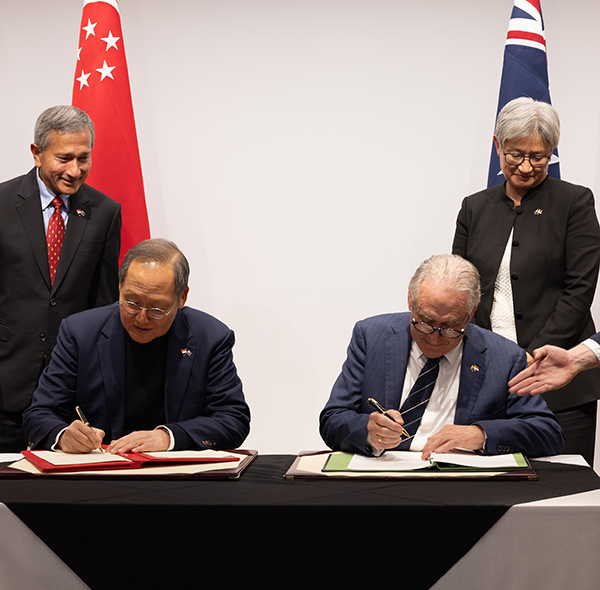 Two officials sign documents at a formal ceremony while other attendees stand behind them. Chinese and Australian flags are displayed in the background.
