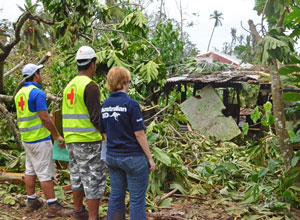Two men in jackets with red cross and a woman in an AusAID shirt inspect cyclone damage to a house; with many trees shredded or blown over