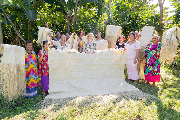 Alt text: Several women wearing traditional colourful Samoan dresses standing on green grass with trees behind them, holding up hand weaved mats.