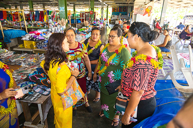 Several women in traditional colourful Samoan dresses wearing flower garlands around their necks, standing in a circle at a market and talking with one another.