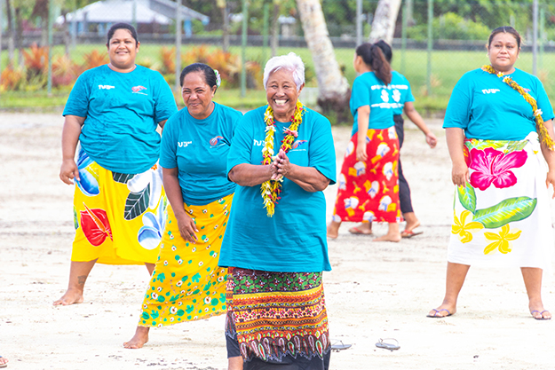 Several women in traditional colourful Samoan dresses, with some wearing flower garlands around their necks, are smiling and playing a sport.  